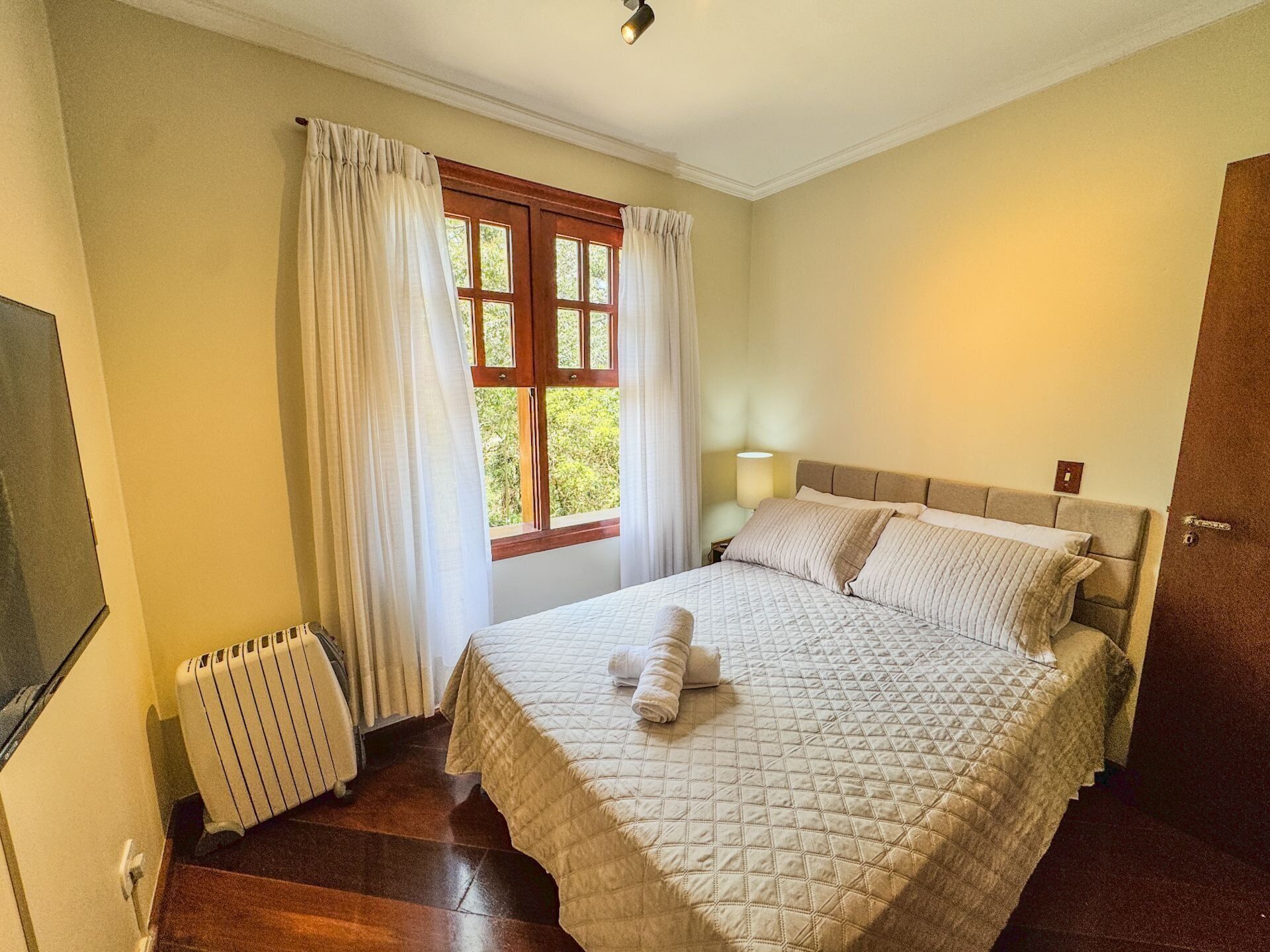 Ein ordentlich eingerichtetes Schlafzimmer mit Bett, Heizkörper, einem Fenster mit Vorhängen und einer Nachttischlampe.