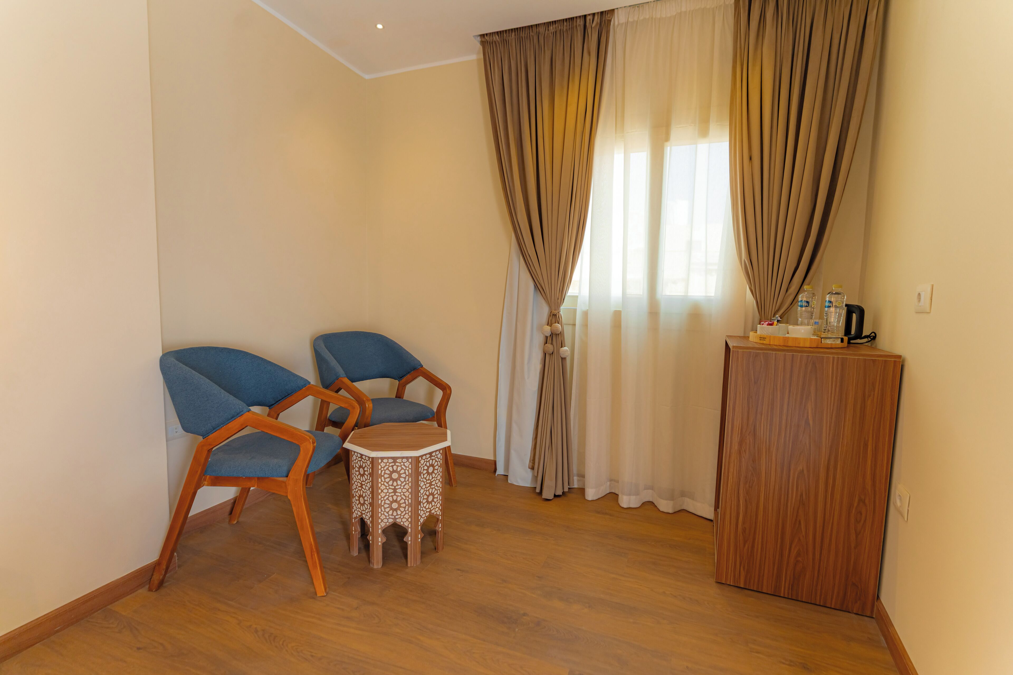 A room with two blue chairs, a small round table, a wooden cabinet, and a window with curtains.