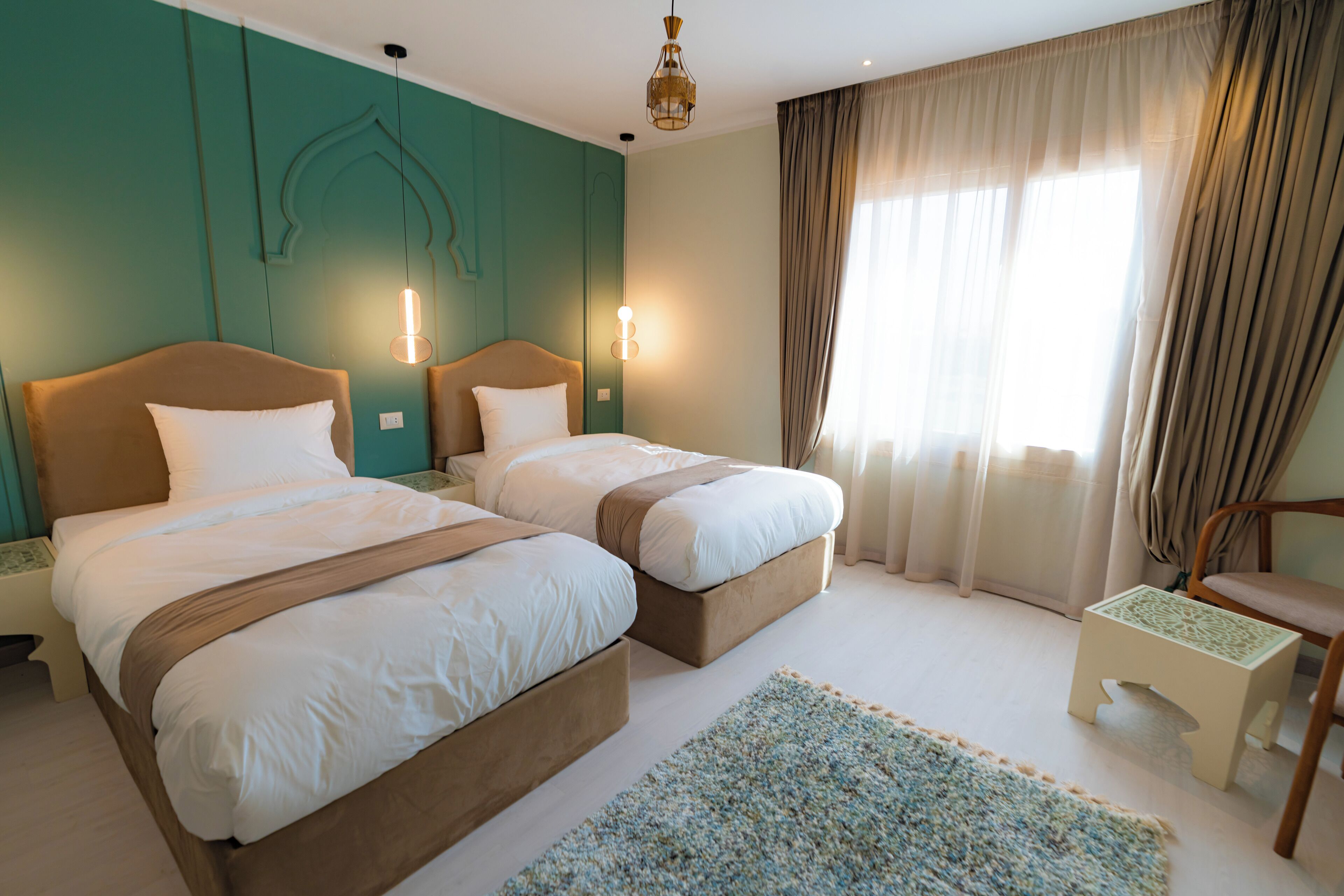 A hotel room with two beds, a small table, a chair, and a window with curtains.