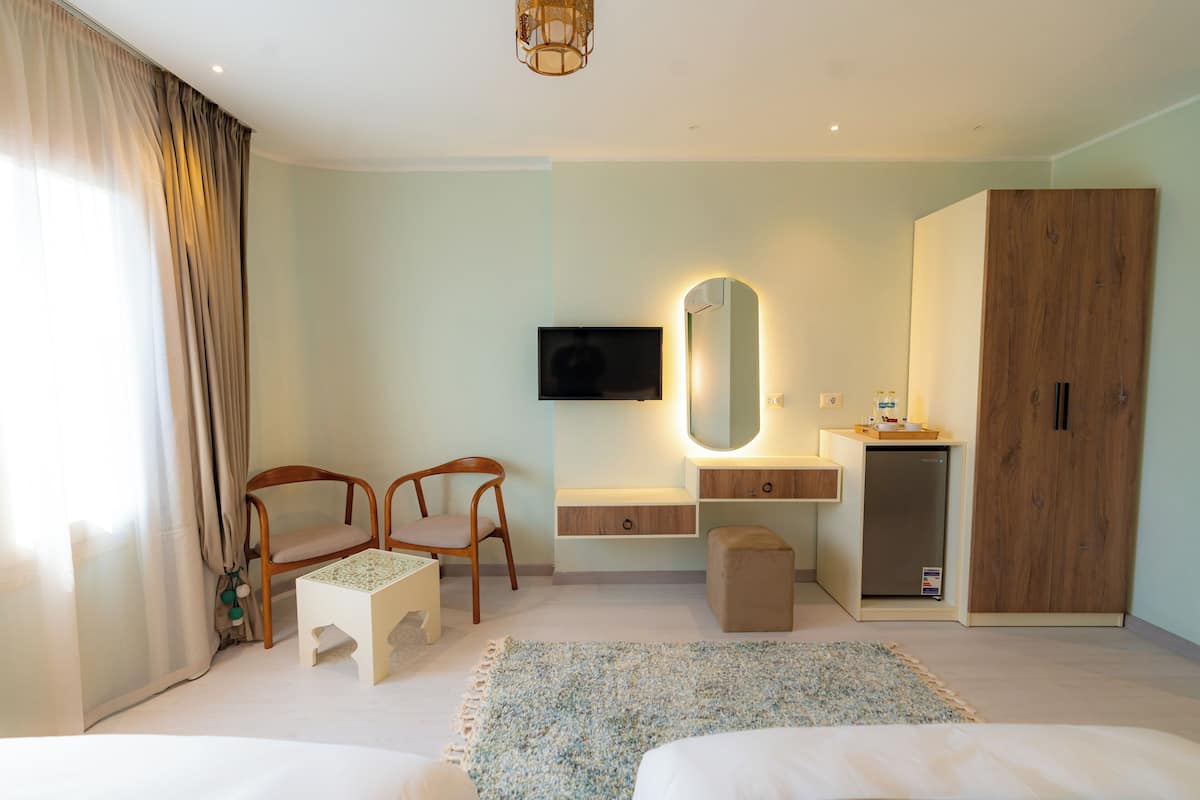 A modern hotel room with a bed, two chairs, a small table, a desk with a mirror, a wardrobe, a television, and a refrigerator.