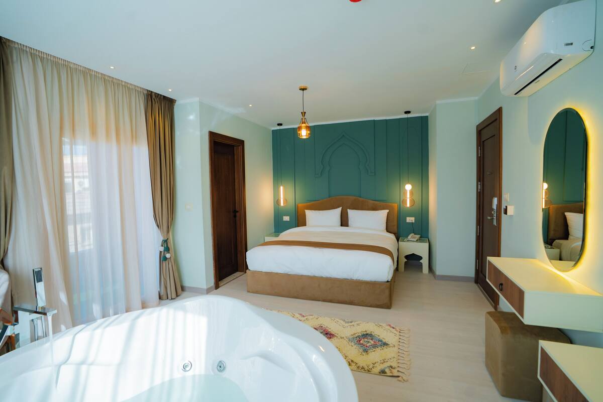 A bedroom with a large bed, a sitting area with a chair and table, a bathtub, and a vanity with a mirror.