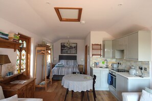 Une chambre compacte comprenant un lit, une table de chevet et un petit bureau avec une chaise. À côté se trouve une kitchenette équipée d’une cuisinière, d’un évier et de placards.