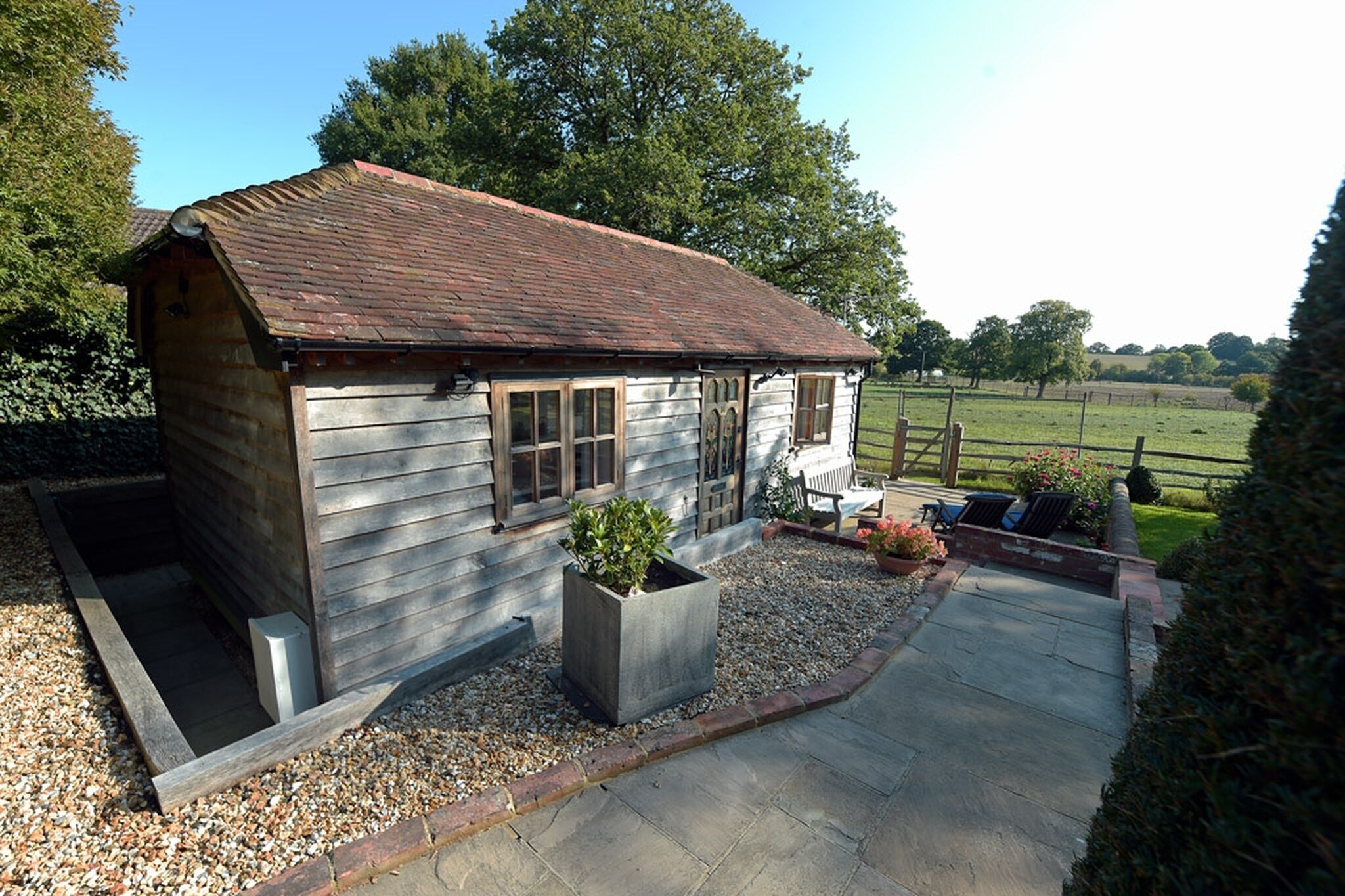 A small wooden cabin with a tiled roof, a gravel driveway, and a garden area with a bench and chairs.