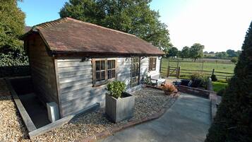 A small wooden cabin with a tiled roof, a gravel driveway, and a garden area with a bench and chairs.