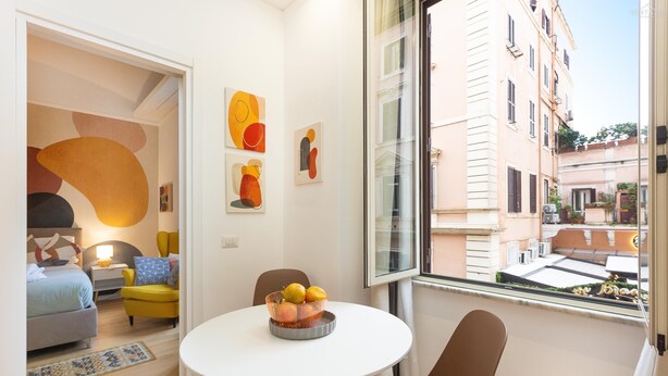 A modern hotel room with a round dining table, a yellow armchair, and a view of the city through a large window. - Rental in Rome Vic House (Rome)