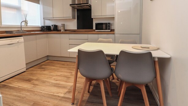 A modern kitchen with white cabinets, a stainless steel range hood, a microwave, and a dining area with two chairs and a table. - Seaside Serenity (Lytham St. Anne's)