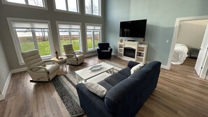 Ein geräumiges Wohnzimmer mit großem Fernseher, bequemen Sitzmöglichkeiten und großen Fenstern mit Blick ins Freie.