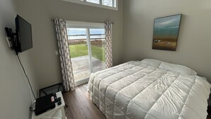 Ett sovrum med en stor säng, en tv som är monterad på väggen, ett skrivbord med en dator och fjärrkontroller samt en skjutdörr i glas som leder ut till en balkong med utsikt över vattnet.