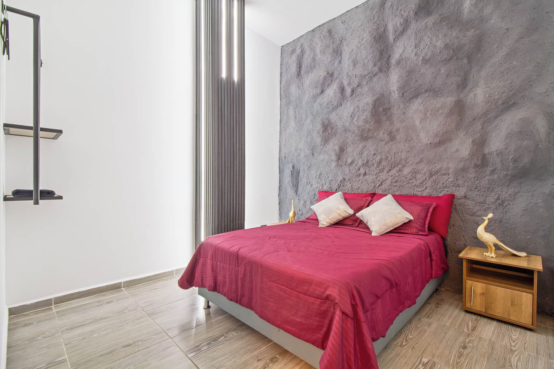 Une chambre moderne avec un mur gris à texture, une couvre-lit rouge et une table de chevet en bois agrémentée d’un objet décoratif.