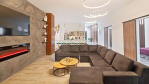 Un salon moderne avec un canapé d’angle, un téléviseur fixé sur un mur à texture, et une cuisine en arrière-plan.