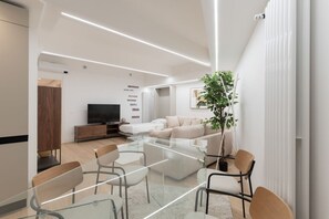 A modern living room with a glass dining table, sofas, a TV, and a plant. - GuestHost - Modern and spacious 96 sqm apartment for 4 guests, located on the semi-basement level of a residential building (stairs to access).The property is just 200 meters from the “De Angeli” M1 Red Line, offering a direct connection to the Duomo (Milan)