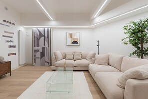 A modern living room with a beige sofa, a glass coffee table, and a large plant. - GuestHost - Modern and spacious 96 sqm apartment for 4 guests, located on the semi-basement level of a residential building (stairs to access).The property is just 200 meters from the “De Angeli” M1 Red Line, offering a direct connection to the Duomo (Milan)