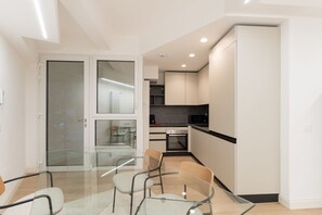 A modern kitchen with white cabinets, a black countertop, and a glass dining table with chairs. - GuestHost - Modern and spacious 96 sqm apartment for 4 guests, located on the semi-basement level of a residential building (stairs to access).The property is just 200 meters from the “De Angeli” M1 Red Line, offering a direct connection to the Duomo (Milan)