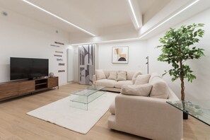 A modern living room with a sofa, TV, glass coffee table, and a potted plant. - GuestHost - Modern and spacious 96 sqm apartment for 4 guests, located on the semi-basement level of a residential building (stairs to access).The property is just 200 meters from the “De Angeli” M1 Red Line, offering a direct connection to the Duomo (Milan)