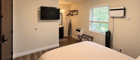 A bedroom with a bed, a TV mounted on the wall, a window with blinds, and a heater on the wall.