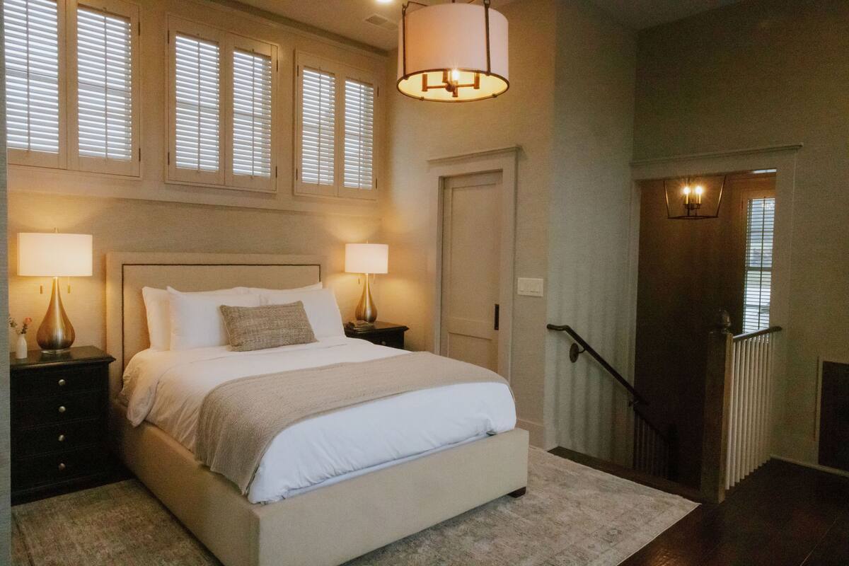 A bedroom with a large bed, two bedside lamps, a window with blinds, a door, and a staircase with a handrail.