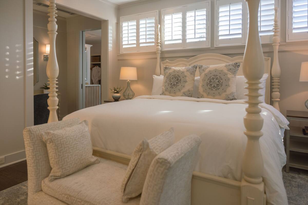A bedroom with a four-poster bed, a bedside table with a lamp, and a chair with cushions.