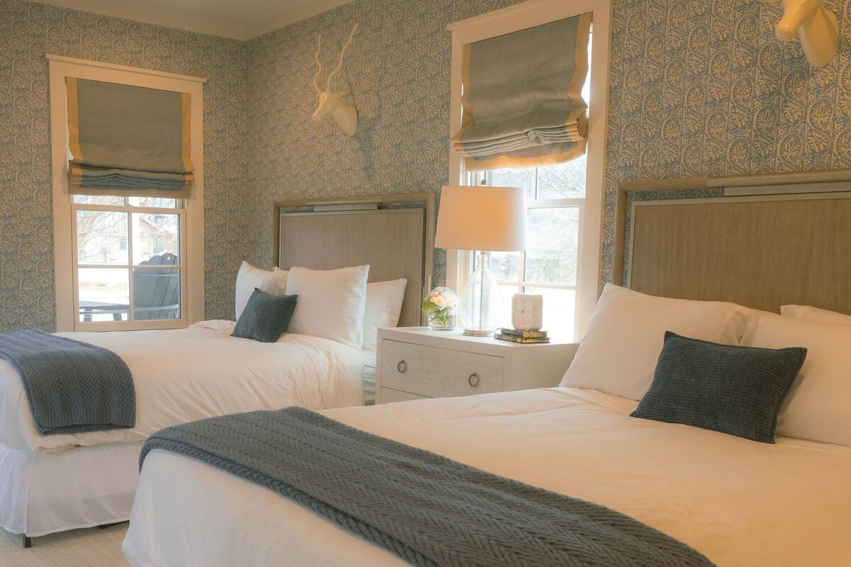A hotel room with two beds, a nightstand, a lamp, and a window with curtains.