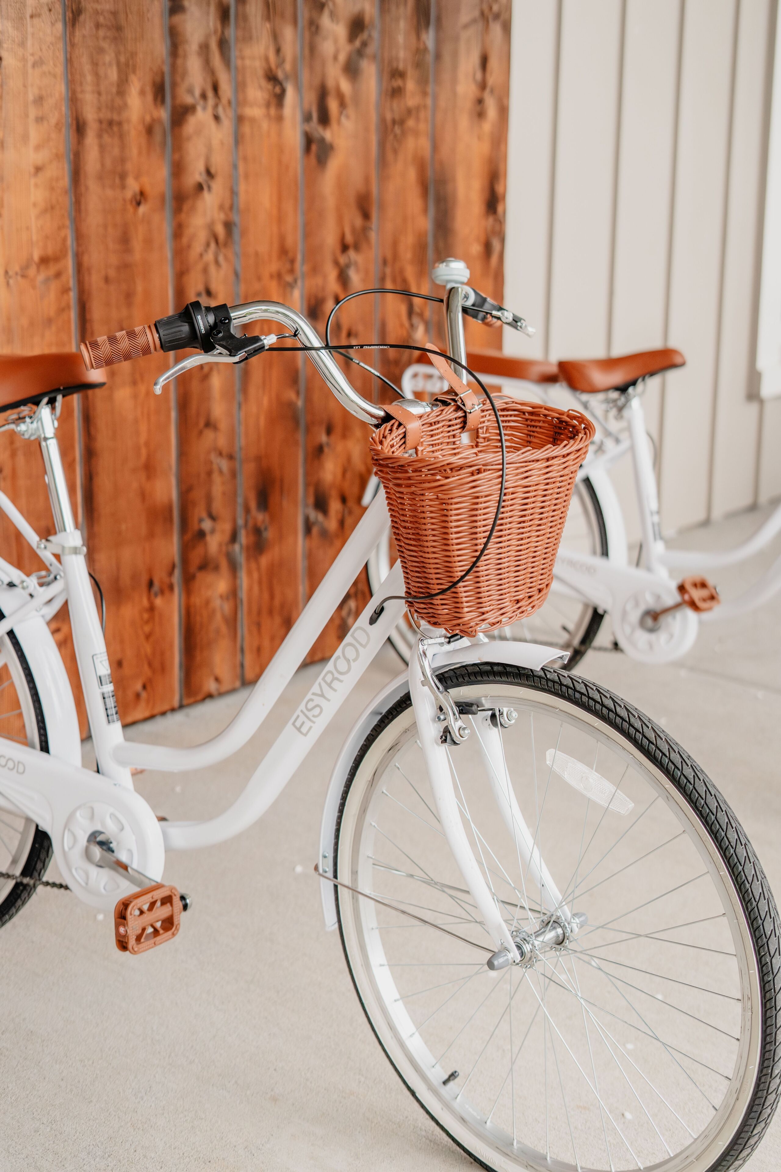 A white bicycle with a wicker basket and brown leather seat, parked against a wooden wall.