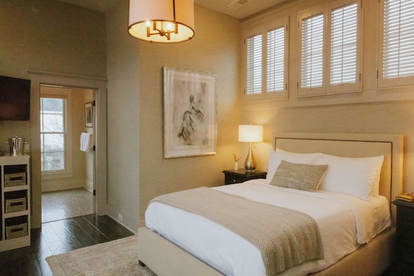 A bedroom with a large bed, a bedside table with a lamp, a framed artwork on the wall, and a window with blinds. - The Inn at The Grove at Whites Creek (Whites Creek)
