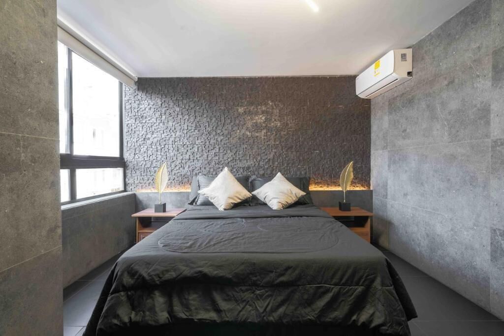 Une chambre moderne avec un mur à la texture de pierre, un grand lit recouvert d’une couverture sombre, deux tables de chevet avec des lampes et un appareil de climatisation fixé au mur.