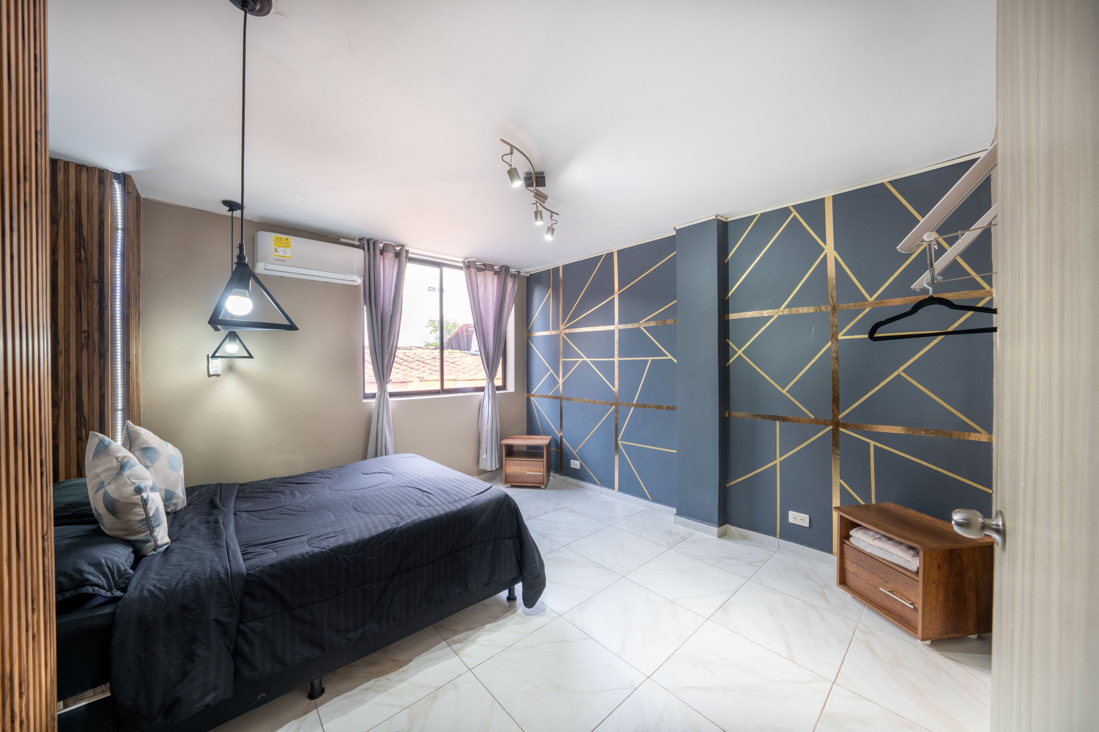 A bedroom with a bed, bedside table, hanging light fixture, and a window with curtains.