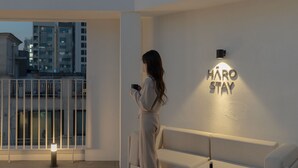 Una persona parada en un balcón con vista a la ciudad, sosteniendo una taza. Un cartel en la pared dice "HARO STAY".