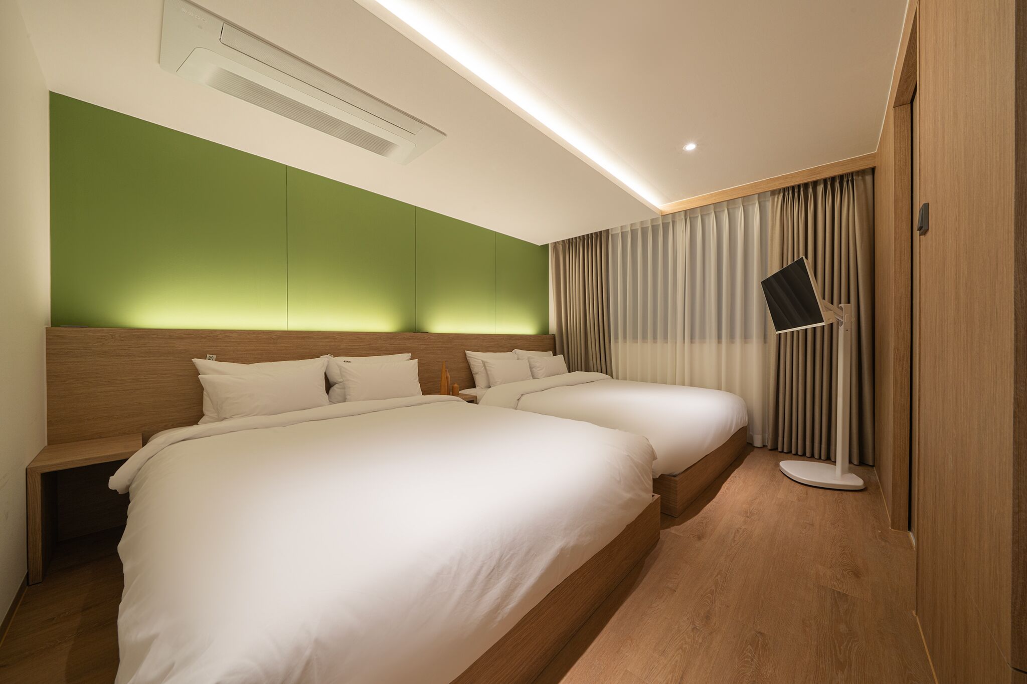 A hotel room with two beds, a green accent wall, a standing desk, and a large window with curtains.