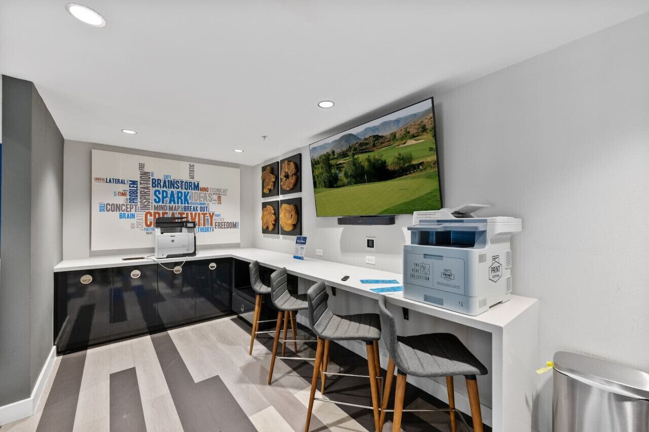 A modern office space with a long white desk, two grey chairs, a large monitor, and a print station.
