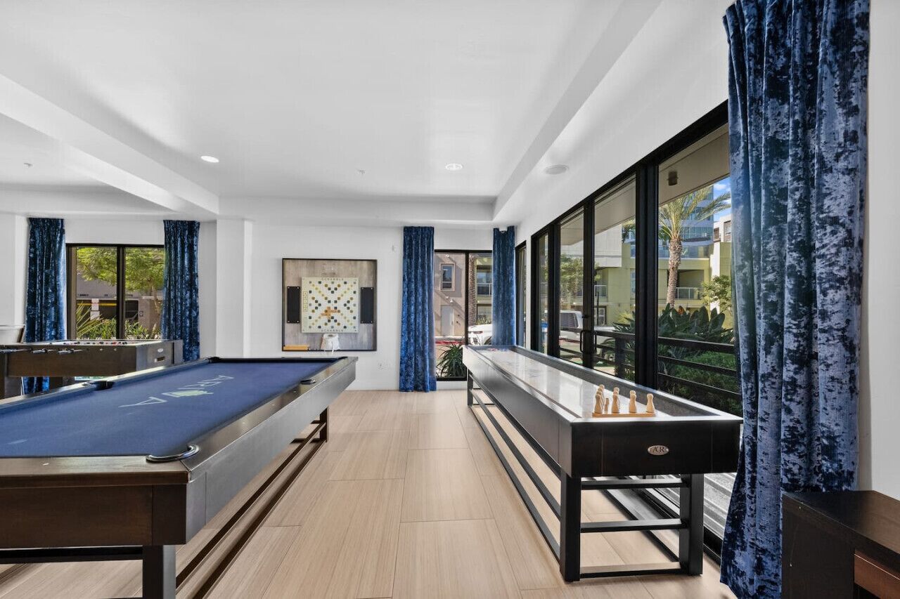 A pool table and a shuffleboard table in a modern room with large windows and a view of the outside.