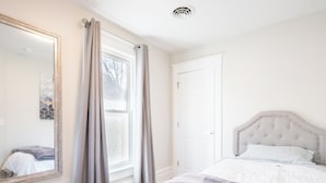 Ein Schlafzimmer mit einem großen Spiegel, einem fenster mit Vorhängen, einem Bett mit einer gesteppten Kopfteil und einer weißen Tür.