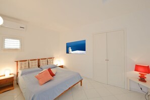 Ein Schlafzimmer mit Bett, Nachttischen, einer Wand-Klimaanlage, einem Fenster mit Jalousien, einer Tür und einem Wandbild.