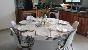 A dining table set for six with white chairs, a vase with flowers, and a kitchen in the background. - Recoin Bohème - Pretty, comfortable accommodation - 5 minutes' walk from the Bassin Bleu! (Saint-Benoît)