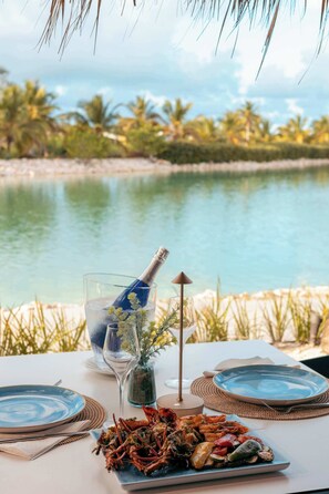 Ein Tisch mit einer Meeresfrüchteplatte, einem Glas Wasser, einer Champagnerflasche und Blick auf einen See mit Palmen.