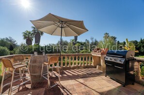 Een terras buiten met een barbecue, stoelen en een tafel.