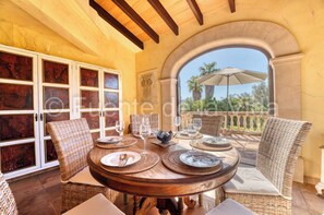 A dining area with a round table set for four, surrounded by wicker chairs, with a view of palm trees through an arched doorway. - Fuenta de la Vida (son servera)