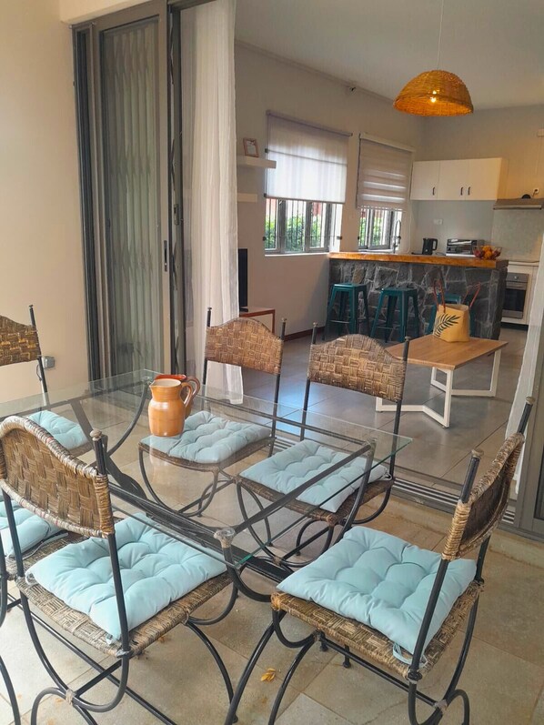 Un coin repas avec des chaises en osier et une table en verre, une cuisine à l’arrière-plan et un mur en pierre.