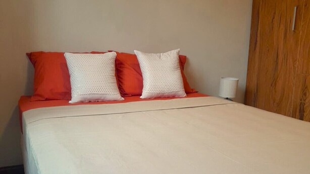 Een bed met een witte dekbedovertrek en twee rode kussens, een houten kledingkast en een witte lamp op een nachtkastje.