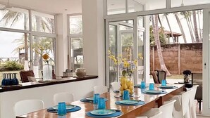 A modern dining area with a wooden table set for six, white chairs, and a large window offering a view of palm trees and a building. - Costa del Sol Beach, El Salvador / Residencia del sol (La Paz)
