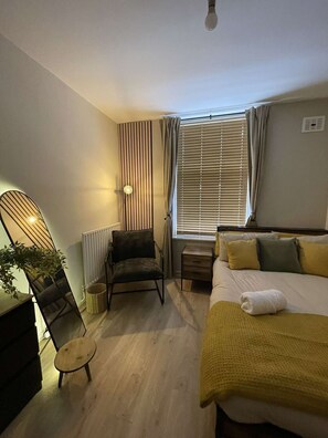 Quarto de hotel pequeno, com cama, poltrona, mesinha, radiador e janela com persianas.