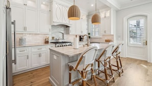 A kitchen with white cabinetry, a central island with bar stools, a stove, and hanging wicker pendant lights. - The Cannon Exchange Building By Charleston Vacays (Charleston)