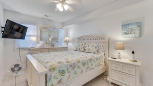 Un dormitorio con cama, mesita de noche, cómoda, ventilador de techo, televisor y ventana con persianas.