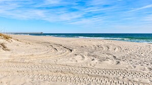 Una playa arenosa con surcos de neumáticos que conducen al océano, un muelle al fondo y un cielo azul despejado.