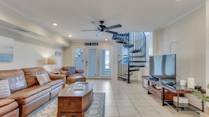 A spacious living room with a leather sofa, a wooden coffee table, a TV, and a spiral staircase leading to a balcony. - Seaview 6&7: 3 Bedroom Duplex in Kure Beach (Kure Beach)