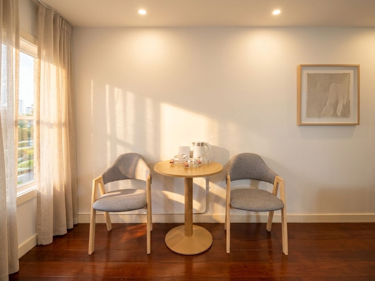 A room with a round wooden table, two grey upholstered chairs, a window with curtains, and a framed artwork on the wall.