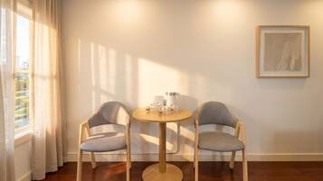 A room with a round wooden table, two grey upholstered chairs, a window with curtains, and a framed artwork on the wall.