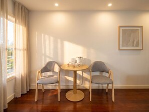 Ein Zimmer mit einem runden Holztisch, zwei grau gepolsterten Stühlen, einem Fenster mit Vorhängen und einem gerahmten Kunstwerk an der Wand.