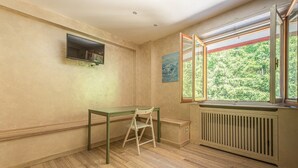 A room with a wooden floor, a table with a chair, a radiator, a window with a view of greenery, and a wall-mounted TV. - La Casina - Pian de` Valli (Pian De` Valli)