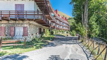 Ein traditionelles Steinhaus mit hölzernen Balkonen, einem Kiesweg und einem bewaldeten Hang im Hintergrund.