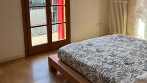 Un dormitorio con cama, suelo de madera, puerta corredera de vidrio, radiador y estante con una planta.